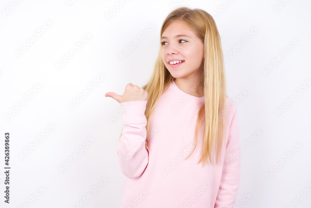 Stupefied beautiful caucasian little girl wearing pink hoodie over white with surprised expression, opens eyes and mouth widely points aside with thumb, shows something strange. Advertisement concept. Stupefied beautiful caucasian little girl wearing pink hoodie over white with surprised expression, opens eyes and mouth widely points aside with thumb, shows something strange. Advertisement concept.