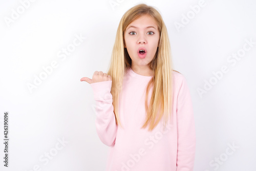 Shocked beautiful caucasian little girl wearing pink hoodie over white background points with thumb away, indicates something. Check this out. Advertisement concept.