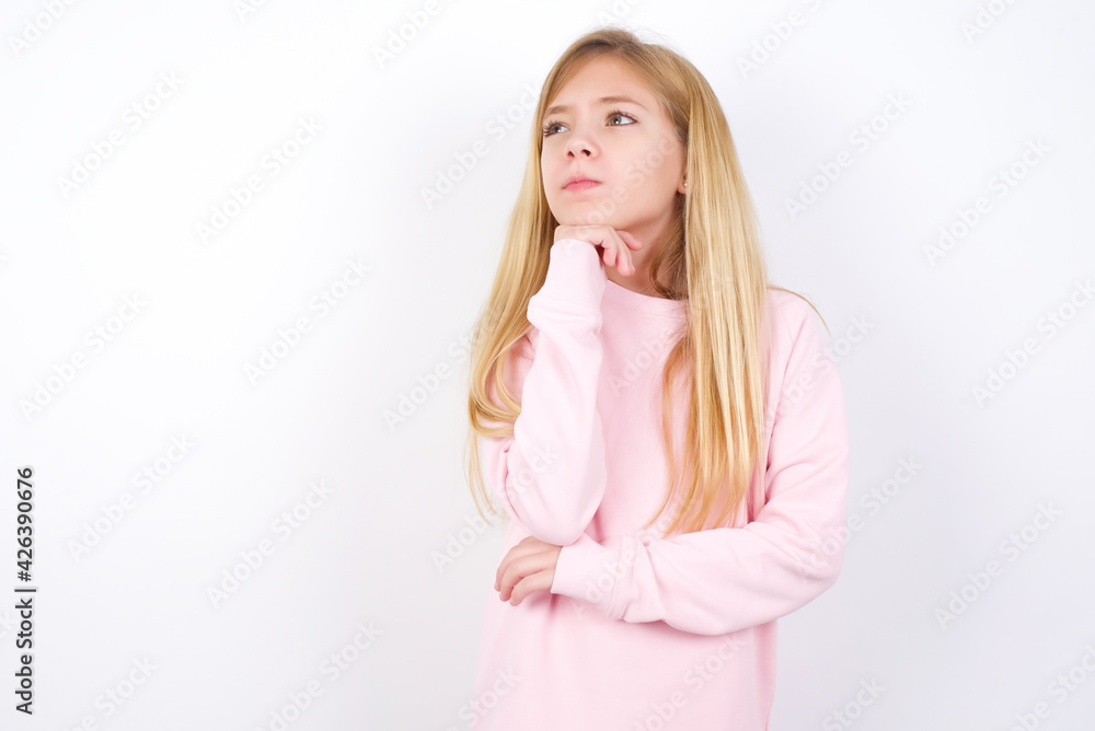 Portrait of thoughtful beautiful caucasian little girl wearing pink hoodie over white keeps hand under chin, looks away trying to remember something or listens something with interest. Youth concept. Portrait of thoughtful beautiful caucasian little girl wearing pink hoodie over white keeps hand under chin, looks away trying to remember something or listens something with interest. Youth concept.