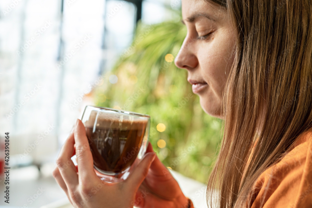 Young pretty Caucasian woman holding glass cup of coffee or cocoa and meditating looking at the cup on background of green house plants, coffeetime, hot drinks, hot chocolate Young pretty Caucasian woman holding glass cup of coffee or cocoa and meditating looking at the cup on background of green house plants, coffeetime, hot drinks, hot chocolate