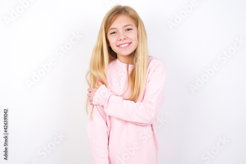 People  lifestyle  youth and happiness concept. Shy pretty beautiful caucasian little girl wearing pink hoodie over white background  feeling happy hugging herself.