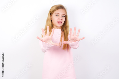 Dissatisfied beautiful caucasian little girl wearing pink hoodie over white background frowns face, has disgusting expression, shows tongue, expresses non compliance, irritated with somebody.