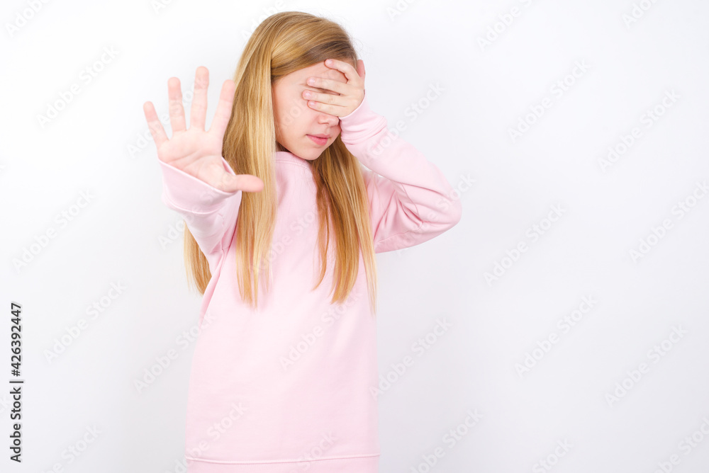 beautiful caucasian little girl wearing pink hoodie over white background covers eyes with palm and doing stop gesture, tries to hide. Don't look at me, I don't want to see, feels ashamed or scared. beautiful caucasian little girl wearing pink hoodie over white background covers eyes with palm and doing stop gesture, tries to hide. Don't look at me, I don't want to see, feels ashamed or scared.