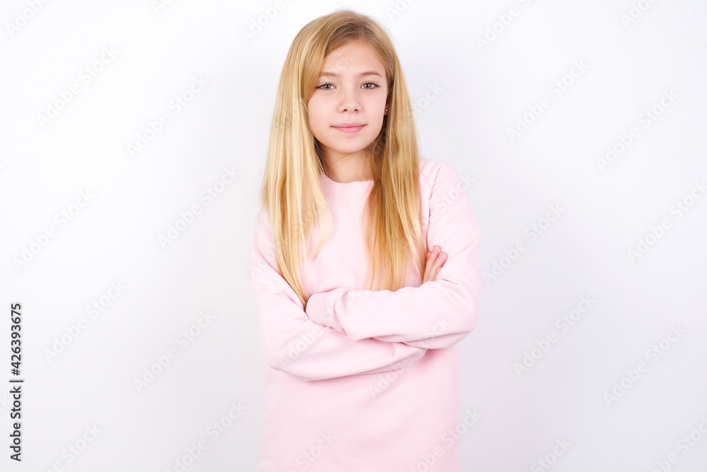 Waist up shot of self confident beautiful caucasian little girl wearing pink hoodie over white background has broad smile, crosses arms, happy to meet with colleagues. Waist up shot of self confident beautiful caucasian little girl wearing pink hoodie over white background has broad smile, crosses arms, happy to meet with colleagues.