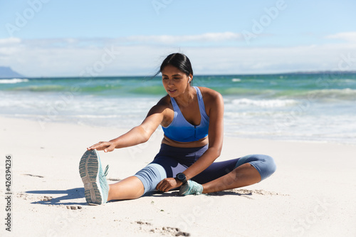 Mixed race woman exercising on beach wearing wireless earphones sitting and stretching