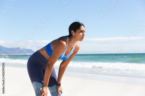 Mixed race woman exercising on beach wearing wireless earphones taking rest