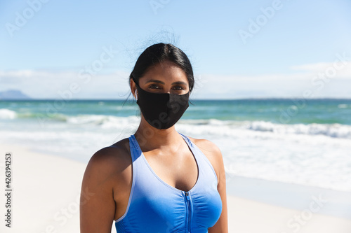 Portrait of mixed race woman exercising on beach wearing face mask and wireless earphones