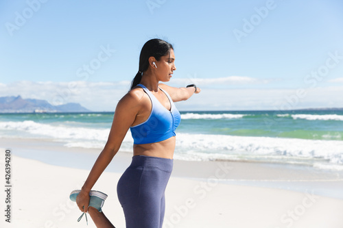 Mixed race woman exercising on beach wearing wireless earphones stretching