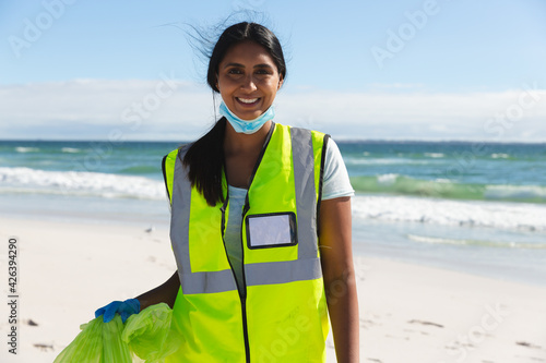 Portrait of happy mixed race woman wearing face mask collecting rubbish from the beach