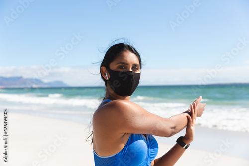 Mixed race woman exercising on beach wearing face mask and wireless earphones stretching arms