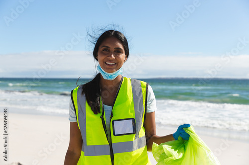 Portrait of smiling mixed race woman wearing face mask collecting rubbish from the beach