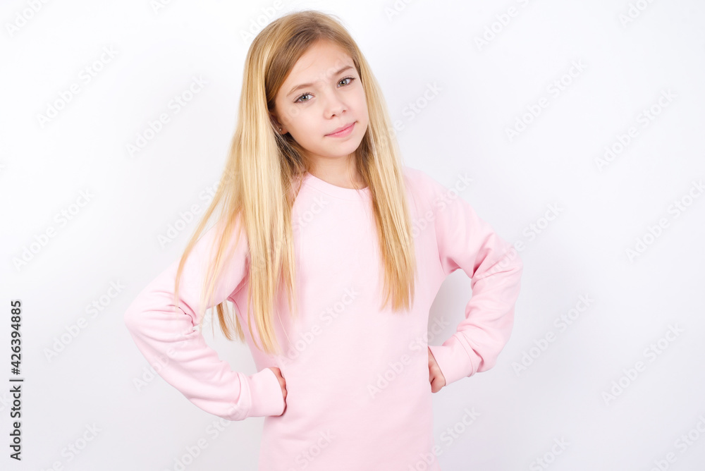 Funny frustrated beautiful caucasian little girl wearing pink hoodie over white background holding hands on waist and silly looking at awkward situation. Funny frustrated beautiful caucasian little girl wearing pink hoodie over white background holding hands on waist and silly looking at awkward situation.