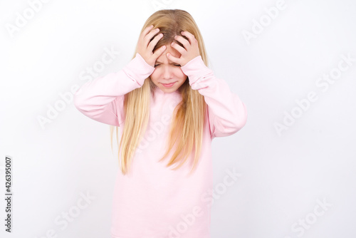 beautiful caucasian little girl wearing pink hoodie over white background holding head with hands, suffering from severe headache, pressing fingers to temples