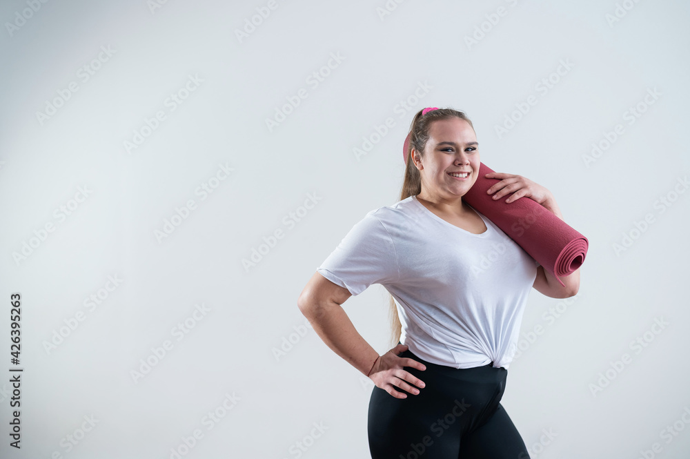 Young fat caucasian woman holding a sport mat. Charming plus size model in sportswear stands on a white background