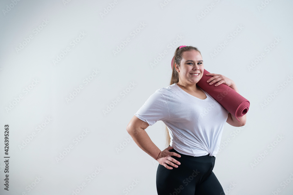 Young fat caucasian woman holding a sport mat. Charming plus size model in sportswear stands on a white background Young fat caucasian woman holding a sport mat. Charming plus size model in sportswear stands on a white background