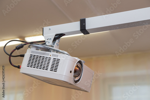 The video projector hangs from the ceiling on a white bracket. The fluorescent lamps are on at the back. Window openings and curtains out of focus. photo