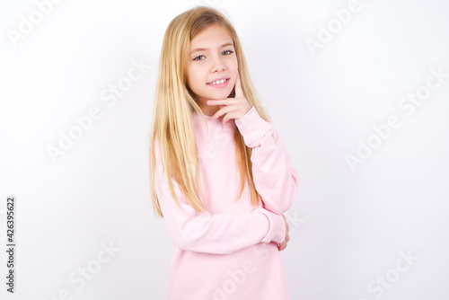 Carefree successful beautiful caucasian little girl wearing pink hoodie over white background touching jawline gazing camera tilting head grinning white teeth delighted. Dental care concept.