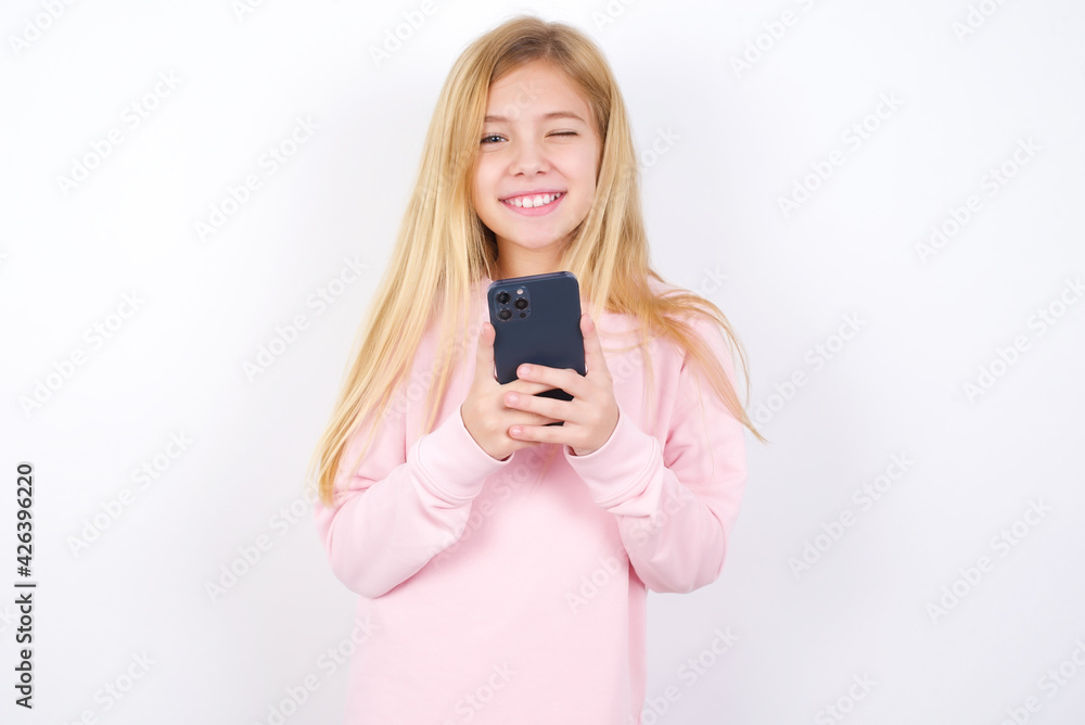 Pleased beautiful caucasian little girl wearing pink hoodie over white background using self phone and looking and winking at the camera. Flirt and coquettish concept. Pleased beautiful caucasian little girl wearing pink hoodie over white background using self phone and looking and winking at the camera. Flirt and coquettish concept.