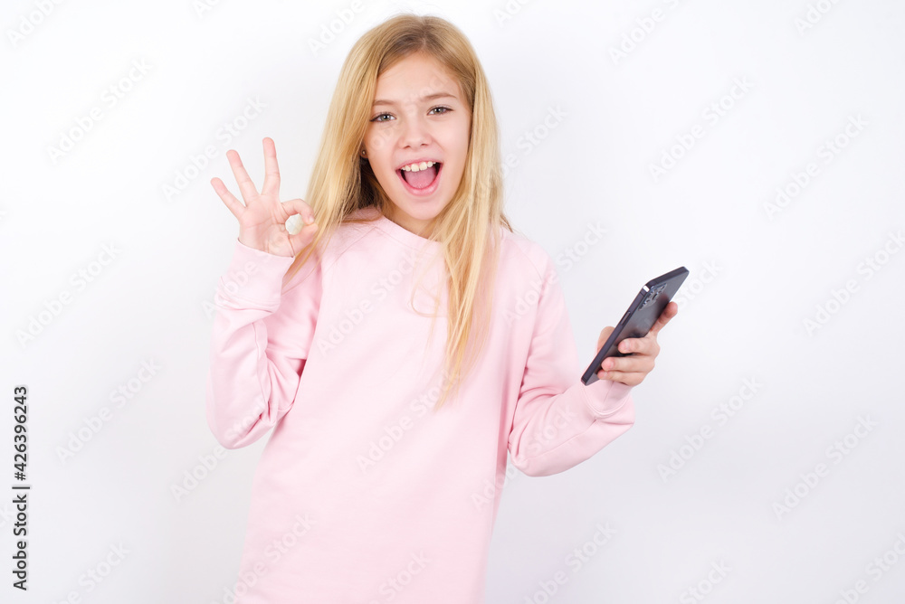 Happy beautiful caucasian little girl wearing pink hoodie over white background sending a message on his smartphone or taking a selfie and making ok sign with his hand. Happy beautiful caucasian little girl wearing pink hoodie over white background sending a message on his smartphone or taking a selfie and making ok sign with his hand.