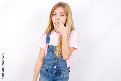 beautiful Caucasian little girl wearing denim overalls over white background covers mouth and looks with wonder at camera, cannot believe unexpected rumors. © Jihan
