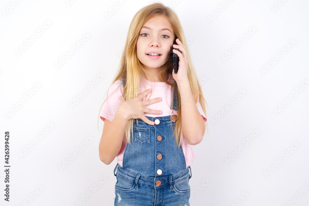 Smiling beautiful Caucasian little girl wearing denim overalls over blue background talks via cellphone, enjoys pleasant great conversation. People, technology, communication concept Smiling beautiful Caucasian little girl wearing denim overalls over blue background talks via cellphone, enjoys pleasant great conversation. People, technology, communication concept