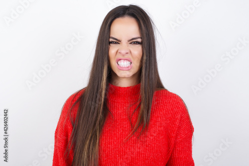 Mad crazy young beautiful brunette woman wearing red knitted sweater over white wall clenches teeth angrily, being annoyed with coming noise. Negative feeling concept. © Jihan