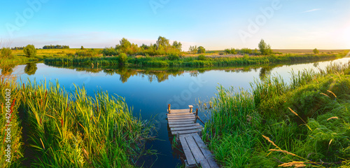Summer sunset landscape with wooden jetty for fishing.Beautiful summertime sunny scene with calm river and green reed.