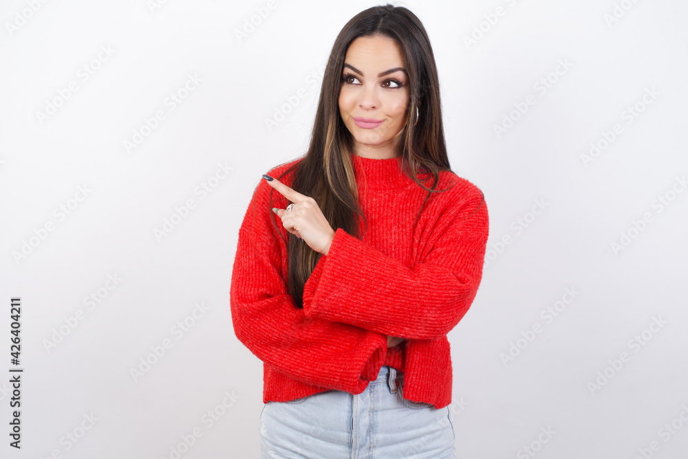 Portrait of young beautiful brunette woman wearing red knitted sweater over white wall posing on camera with tricky look, presenting product with index finger. Advertisement concept. Portrait of young beautiful brunette woman wearing red knitted sweater over white wall posing on camera with tricky look, presenting product with index finger. Advertisement concept.