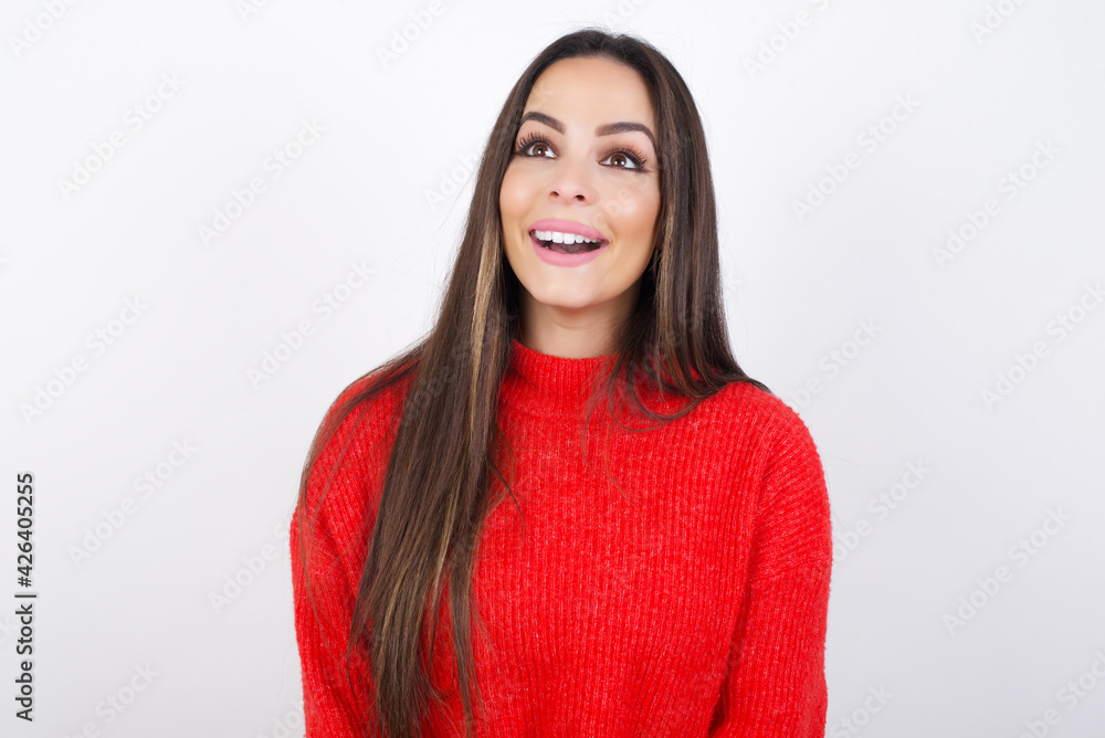 Surprised young beautiful brunette woman wearing red knitted sweater over white wall, shrugs shoulders, looking sideways, being happy and excited. Sudden reactions concept. Surprised young beautiful brunette woman wearing red knitted sweater over white wall, shrugs shoulders, looking sideways, being happy and excited. Sudden reactions concept.