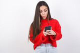 young beautiful brunette woman wearing red knitted sweater over white wall looks with bugged eyes, holds modern smart phone, receives unexpected message from friend, reads reminder.