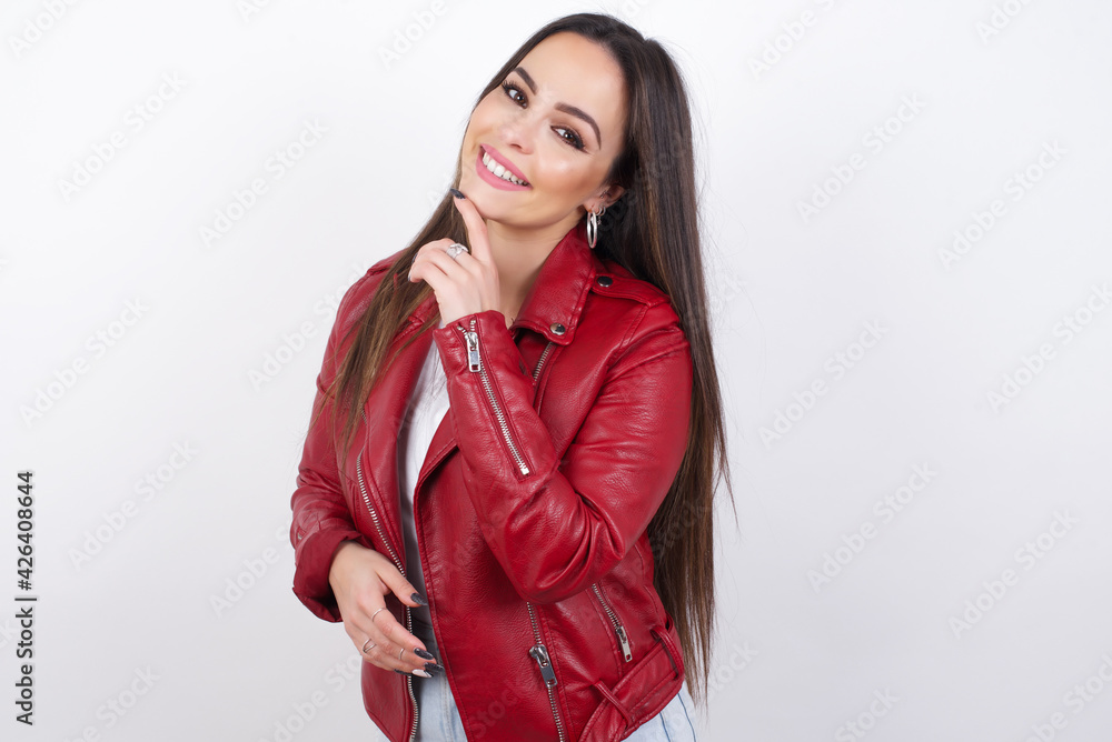 young beautiful brunette woman wearing red biker jacket over white wall laughs happily keeps hand on chin expresses positive emotions smiles broadly has carefree expression young beautiful brunette woman wearing red biker jacket over white wall laughs happily keeps hand on chin expresses positive emotions smiles broadly has carefree expression