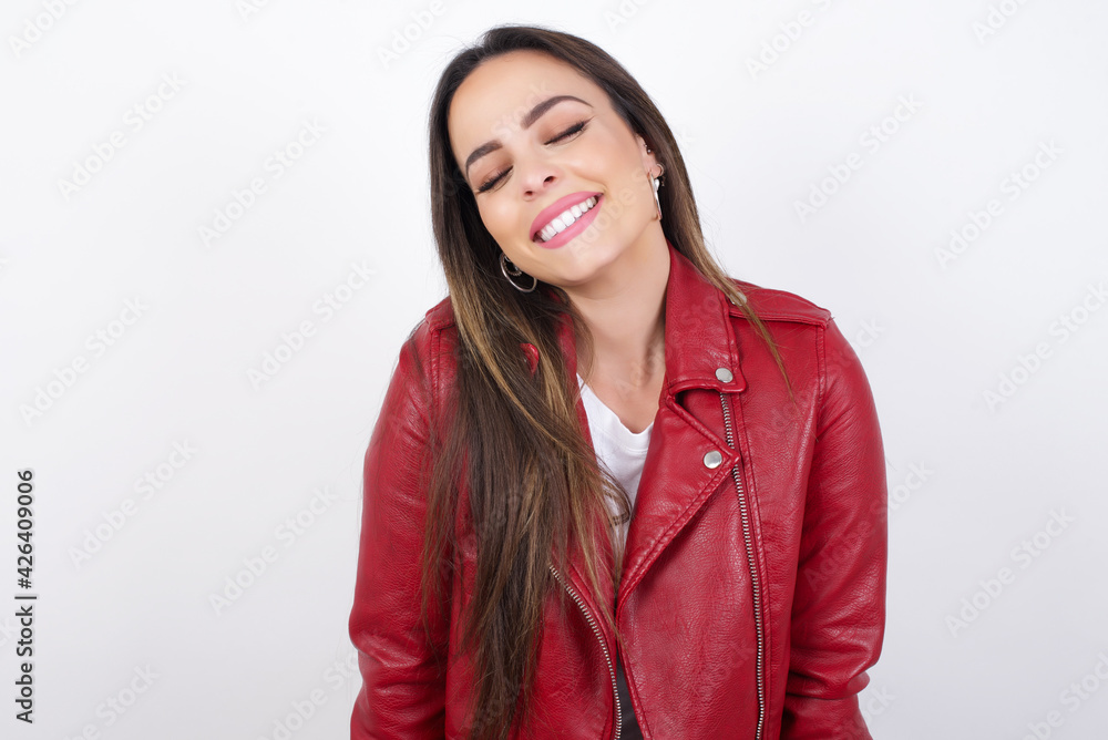 Positive young beautiful brunette woman wearing red biker jacket over white wall with overjoyed expression closes eyes and laughs shows white perfect teeth Positive young beautiful brunette woman wearing red biker jacket over white wall with overjoyed expression closes eyes and laughs shows white perfect teeth