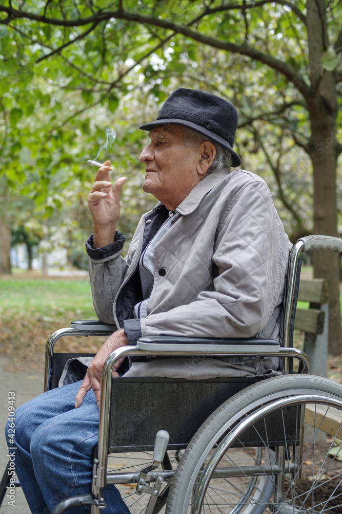 Disabled sad senior man smokes in outdoor while sitting in wheelchair Disabled sad senior man smokes in outdoor while sitting in wheelchair