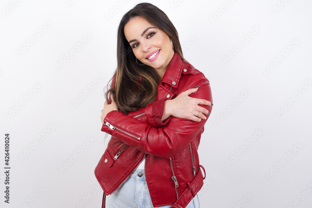 Charming pleased young beautiful brunette woman wearing red biker jacket over white wall embraces own body, pleasantly feels comfortable poses. Tenderness and self esteem concept Charming pleased young beautiful brunette woman wearing red biker jacket over white wall embraces own body, pleasantly feels comfortable poses. Tenderness and self esteem concept