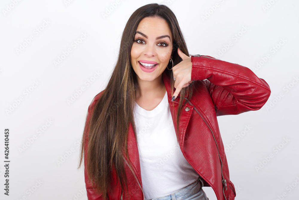 young beautiful brunette woman wearing red biker jacket over white wall makes phone gesture, says call me back again, has glad expression. young beautiful brunette woman wearing red biker jacket over white wall makes phone gesture, says call me back again, has glad expression.
