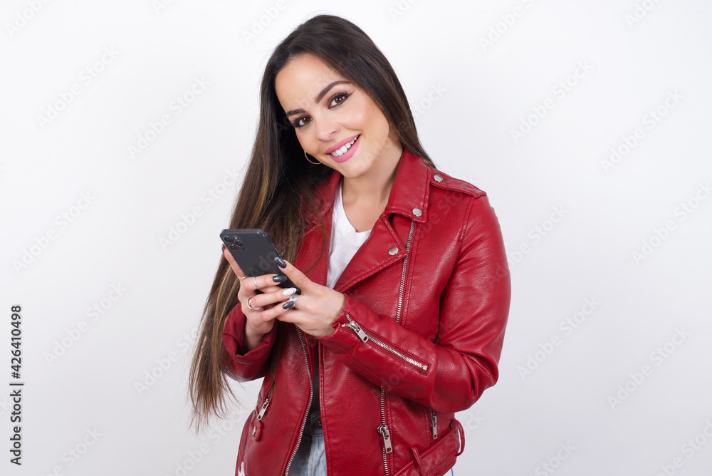young beautiful brunette woman wearing red biker jacket over white wall Mock up copy space. Using mobile phone, typing sms message young beautiful brunette woman wearing red biker jacket over white wall Mock up copy space. Using mobile phone, typing sms message