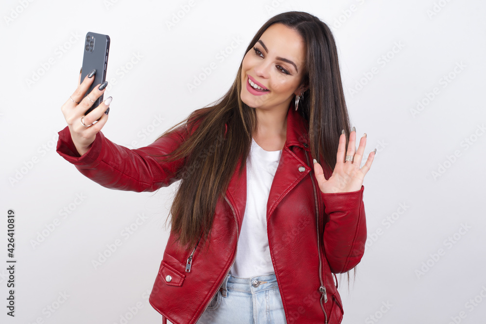 Naklejka premium young beautiful brunette woman wearing red biker jacket over white wall holds modern mobile phone and makes video call waves palm in hello gesture. People modern technology concept