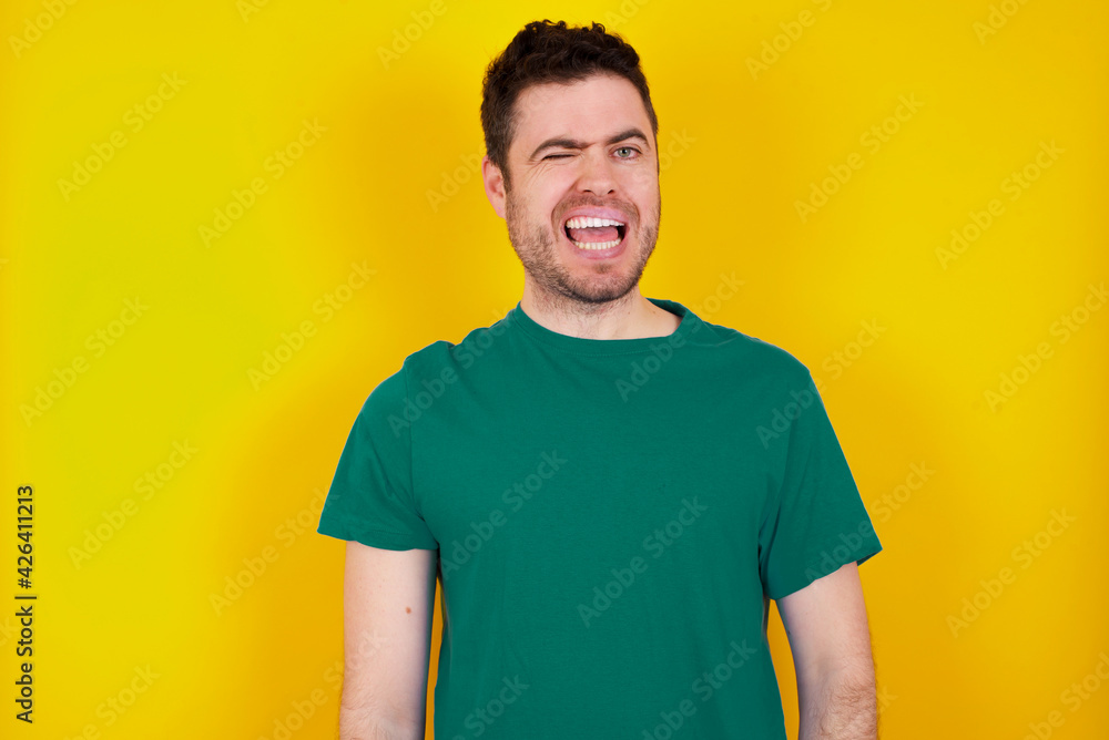 Coquettish young Caucasian man wearing green T-shirt against yellow wall smiling happily, blinking at camera in a playful manner, flirting with you. Coquettish young Caucasian man wearing green T-shirt against yellow wall smiling happily, blinking at camera in a playful manner, flirting with you.