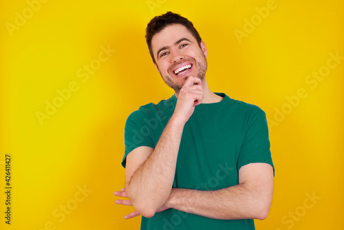 Photography Optimistic young Caucasian man wearing green T-shirt against yellow wall keeps hands partly crossed and hand under chin, looks at camera with pleasure