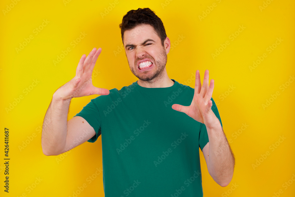 young Caucasian man wearing green T-shirt against yellow wall Shouting frustrated with rage, hands trying to strangle, yelling mad. young Caucasian man wearing green T-shirt against yellow wall Shouting frustrated with rage, hands trying to strangle, yelling mad.
