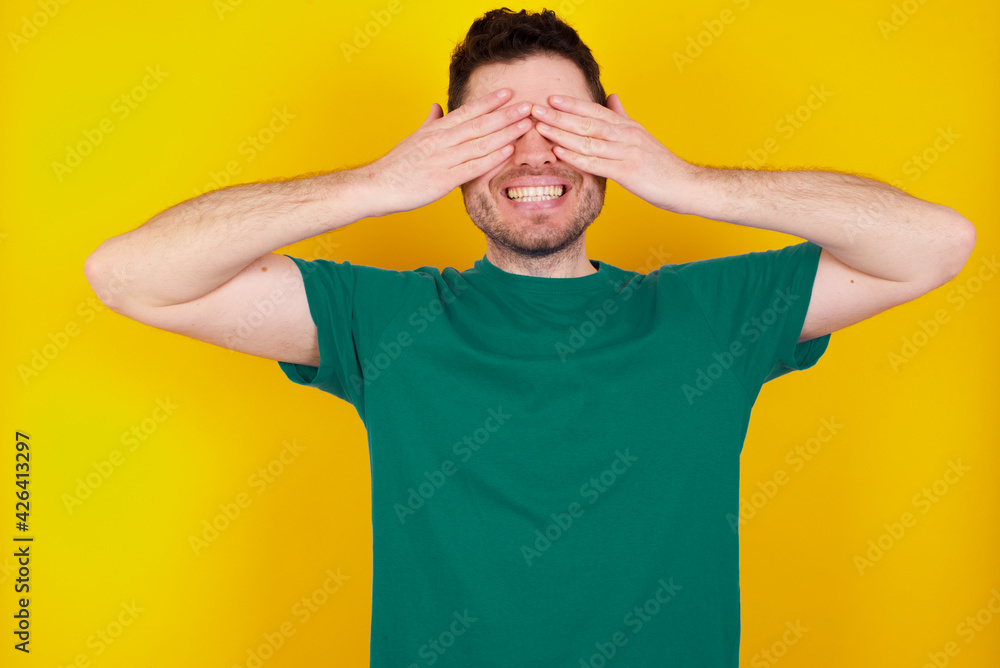 young Caucasian man wearing green T-shirt against yellow wall covering eyes with hands smiling cheerful and funny. Blind concept.