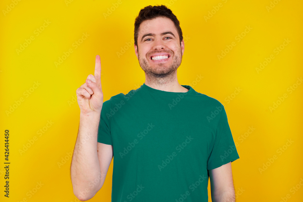 young Caucasian man wearing green T-shirt against yellow wall showing and pointing up with finger number one while smiling confident and happy. young Caucasian man wearing green T-shirt against yellow wall showing and pointing up with finger number one while smiling confident and happy.