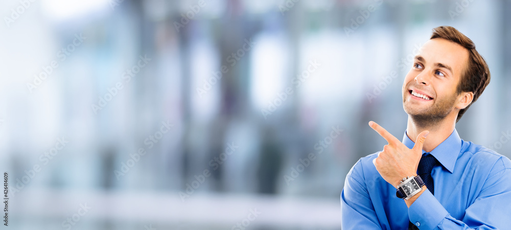 Looking up, smiling businessman pointing showing something, over blurred modern office interior background. Business, job and education coach concept. Confident man pointing at copy space. Looking up, smiling businessman pointing showing something, over blurred modern office interior background. Business, job and education coach concept. Confident man pointing at copy space.