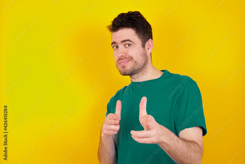 Joyful young Caucasian man wearing green T-shirt against yellow wall wink and points index fingers at camera, chooses someone, makes finger gun pistol. Joyful young Caucasian man wearing green T-shirt against yellow wall wink and points index fingers at camera, chooses someone, makes finger gun pistol.