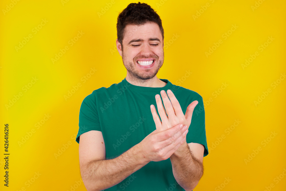 young Caucasian man wearing green T-shirt against yellow wall Suffering pain on hands and fingers, arthritis inflammation young Caucasian man wearing green T-shirt against yellow wall Suffering pain on hands and fingers, arthritis inflammation