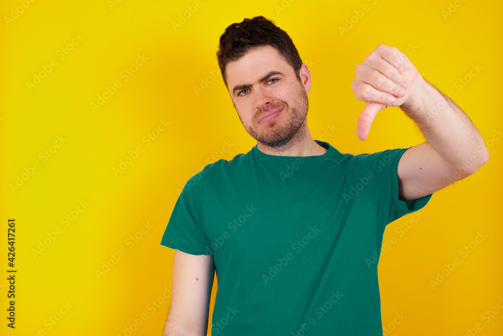 young Caucasian man wearing green T-shirt against yellow wall feeling angry, annoyed, disappointed or displeased, showing thumbs down with a serious look young Caucasian man wearing green T-shirt against yellow wall feeling angry, annoyed, disappointed or displeased, showing thumbs down with a serious look