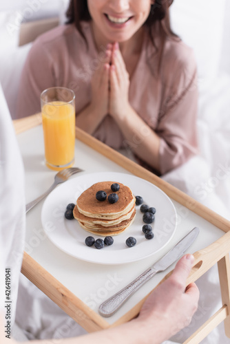 Cropped view of man holding breakfast tray with pancakes and berries near smiling wife on blurred background on bed