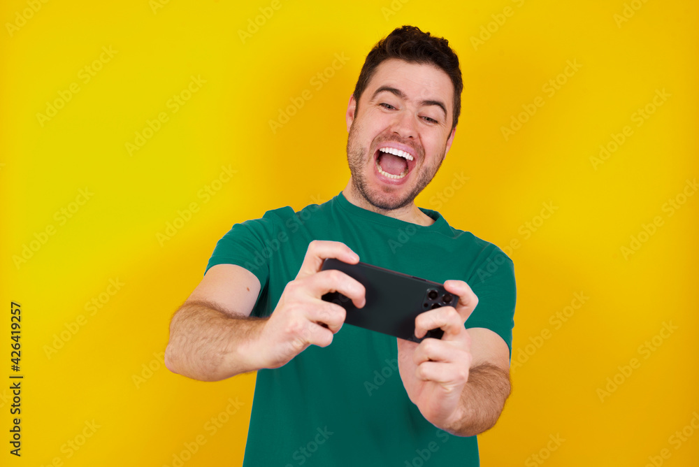 Crazy impressed young handsome caucasian man wearing green t-shirt against yellow background use cell phone reads incredible news on social media information stare, screams wow omg. Crazy impressed young handsome caucasian man wearing green t-shirt against yellow background use cell phone reads incredible news on social media information stare, screams wow omg.