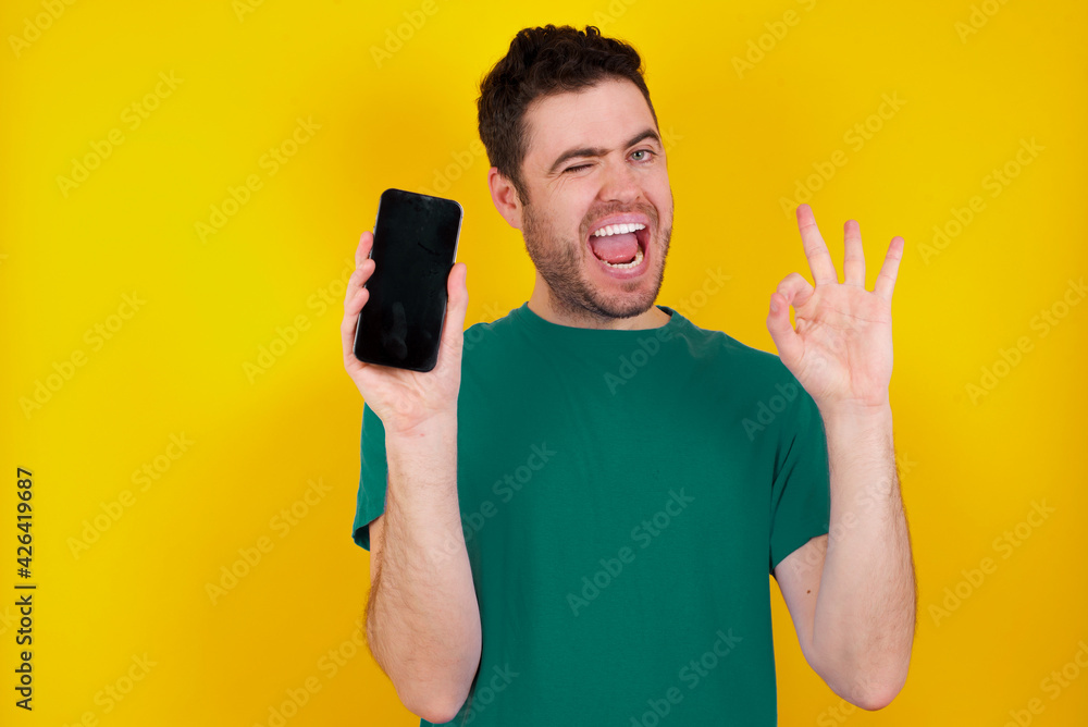 young handsome caucasian man wearing green t-shirt against yellow background holding in hands cell showing ok-sign young handsome caucasian man wearing green t-shirt against yellow background holding in hands cell showing ok-sign