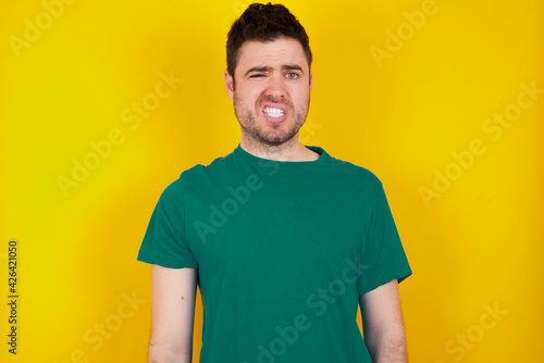 Portrait of dissatisfied young handsome caucasian man wearing green t-shirt against yellow wall smirks face, purses lips and looks with annoyance at camera, discontent hearing something unpleasant